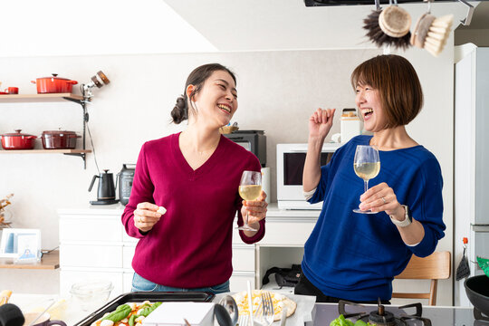 Two cheerful Asian women laughing heartily and holding wine glasses in a modern kitchen