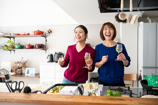 Two happy Asian women holding wine glasses and dancing playfully while making homemade pizzas in a bright kitchen