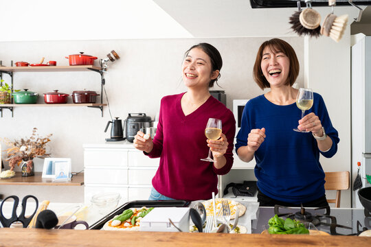 Two happy Asian women holding wine glasses and dancing playfully while making homemade pizzas in a bright kitchen