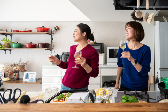 Two smiling Asian women holding wine glasses and looking away together in a bright kitchen