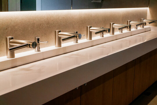 Row of modern automatic sensor water taps integrated with hand dryers over a long white corian sink in a public restroom