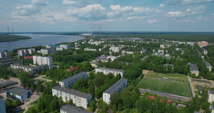 Aerial orbiting shot of a green provincial town featuring a football pitch, residential neighborhoods, and a large power plant in the distance