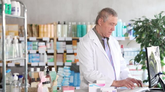 Confident professional senior pharmacist in white coat working behind drugstore counter, inputting purchase orders into computer system, managing incoming supplies and ensuring stock availability
