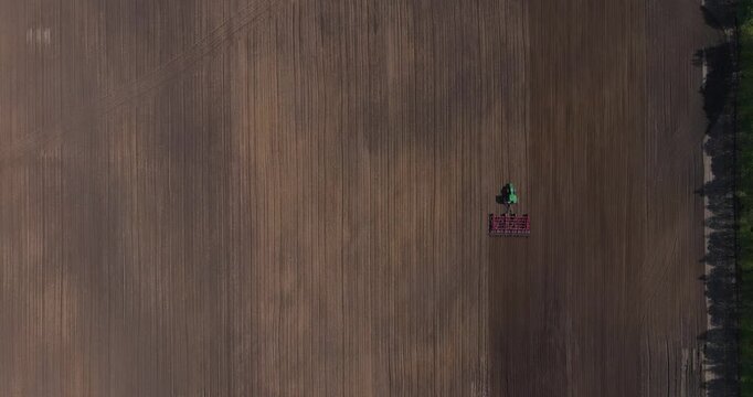 Top-down aerial view directly above an agricultural tractor pulling a red harrow disc plow on a large brown soil field