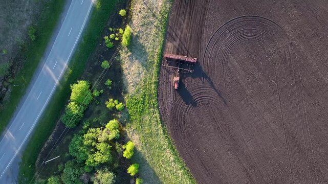 Tractor with seed drill working along edge of agricultural field near rural road and forest belt aerial drone view spring farming operations and countryside landscape
