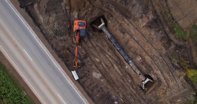 Top-down aerial view of an orange excavator laying a large black pipe into a deep trench beside a road