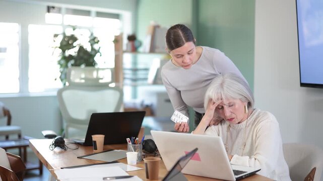 Adult woman sympathizes to sick senior female manager with headache and offers pain pills. Co-worker helps elderly woman suffering from headache. She offers help by participating in solving problem