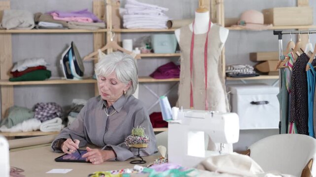 Mature dressmaker draws a sketch of clothes and patterns on an electronic tablet. Woman creates a fashion design in a workshop.