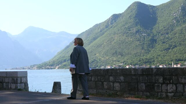 Mature Woman Walking by Mountain Bay. Mature woman walking along a quiet waterfront road with green mountains and a calm bay in the background. Coastal travel and peaceful outdoor lifestyle scene.