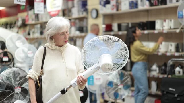 Senior woman chooses a fan for the summer heat. Elderly woman stands in a home appliance store and buys a fan with different airflow modes for living room. High quality 4k footage