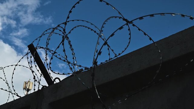 Barbed wire fencing against a backdrop of blue sky and clouds, illustrating a boundary or security feature. The sharp edges of the wire stand out, emphasizing the purpose of protection