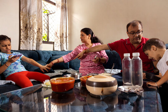 father asking his son to sit on sofa for lunch while mother is scolding here daughter to put phone down for eating lunch