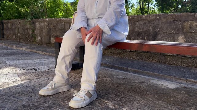 Senior Woman Holding Knee While Sitting on Bench. Close-up of a senior woman sitting on an outdoor bench and holding her knee with both hands. Joint pain, knee discomfort and mobility problem concept.