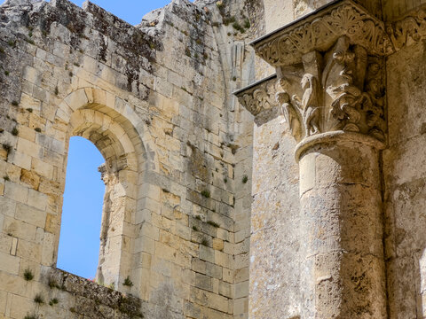 Visitors walk around the historic Abbaye de la Sauve-Majeur in Gironde, France. The ruins stand under a blue sky with clouds, revealing details of the architecture and scenery.