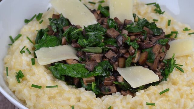 A bowl contains a serving of mushroom and spinach risotto. On top are pieces of Morteau sausage and parmesan cheese. The dish is placed on a wooden surface.