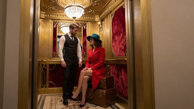 A fashionable woman in a red coat and blue hat sits atop vintage luggage inside a luxurious, gold-trimmed elevator with a porter standing nearby.