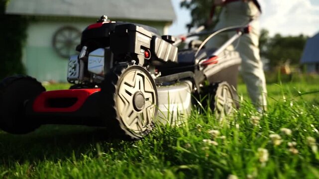 Lawn mower moves through vibrant green grass. Operator guides machine with focused, steady motion. Sunlight glints off blades as they cut tall grass. Perfect for home improvement or gardening projects