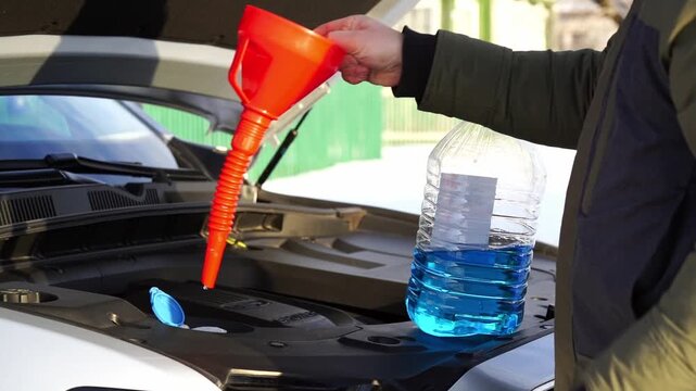 Person pours coolant into engine with funnel. Hands steady as blue fluid flows into car's system. Engine bay open, revealing inner workings. Scene implies routine maintenance or emergency fix