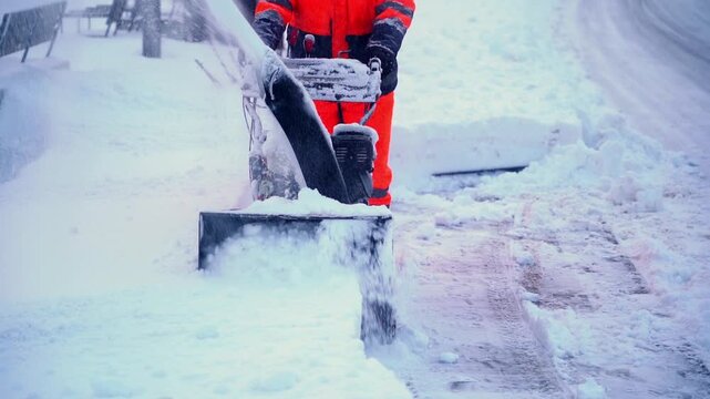 Worker clears snow with snow blower. Snow sprays from machine as it moves forward. Worker wears bright orange safety gear. Scene shows cold winter street cleanup. Ideal for weather, winter