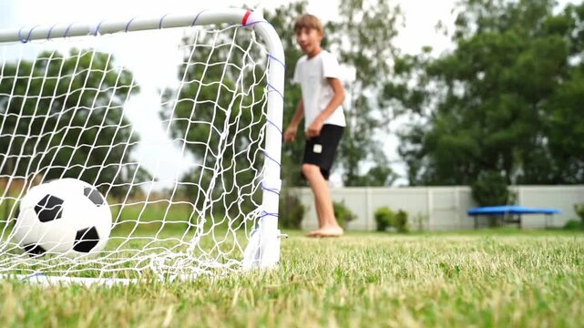 Young boy runs toward soccer ball near goalpost. Grass is green, sunlit, and slightly uneven. Background trees sway gently in breeze. He wears simple white shirt and dark shorts
