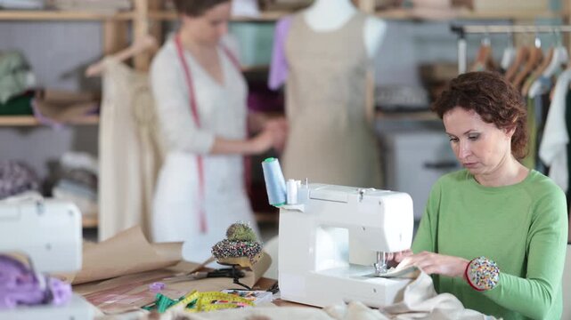 Woman designer sews clothes using sewing machine, perform edge finishing stitching seam. Companion work in background with mannequin