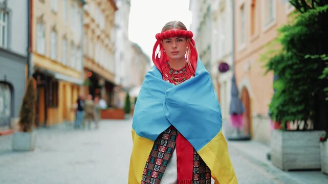 Confident woman dressed in traditional Ukrainian vyshyvanka, red floral wreath, and flag walking along a historic cobblestone street. Urban background with buildings.