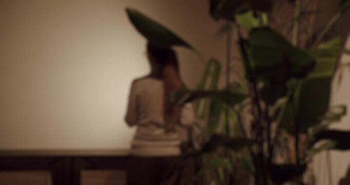 Silhouette of an unrecognizable woman with a ponytail walking in slow motion inside a dimly lit room, with a large green houseplant creating a mysterious and atmospheric foreground