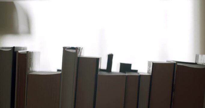 Unrecognizable person walking in the background of a bookshelf with numerous silhouetted books against a bright window, creating a serene and thoughtful atmosphere in a quiet library or study