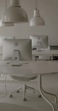 Modern, monochromatic white office interior showing empty chairs and desks with computers under industrial-style lamps. Clean and minimalist coworking space ready for business without people. vertical