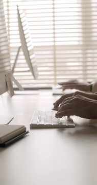 Close-up side view of a person's hands typing on a wireless keyboard at a minimalist office desk, with another person working on a computer in the blurred background near a window. vertical