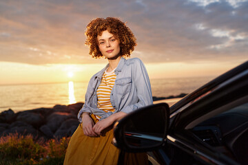 Fototapeta premium Young woman with curly hair leans on a car by a rocky shore at sunset, wearing a denim jacket over a striped top and orange skirt, radiating relaxed confidence