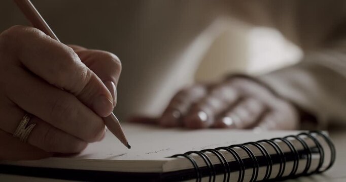 Slow motion, close-up shot of a woman's hands carefully writing or drawing with a graphite pencil in a spiral-bound notepad, expressing creativity and concentration on her art