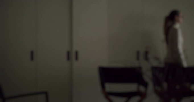 Defocused shot of a lonely woman with a ponytail walking away from a bright light source and leaving a dark, modern apartment, symbolizing sadness, memory, or a mysterious departure