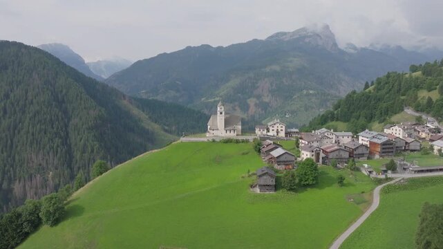 Aerial view of Colle Santa Lucia village and historic church overlooking a green alpine hillside in the Dolomites. Scenic mountain landscape with traditional architecture and dramatic peaks.
