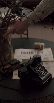 Slow motion footage of a vintage black rotary phone, newspaper, and decor on a dark coffee table with a woman's hand touching the flowers in a vase in a cozy, moody interior living room. vertical