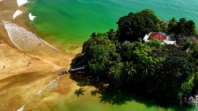 Small forested island with a red-roof temple sits beside a sandy beach and shallow green sea as the camera flies overhead.