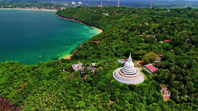 Japanese Peace Pagoda rises from dense Rumassala forest as a drone flies over the curved bay and turquoise sea.