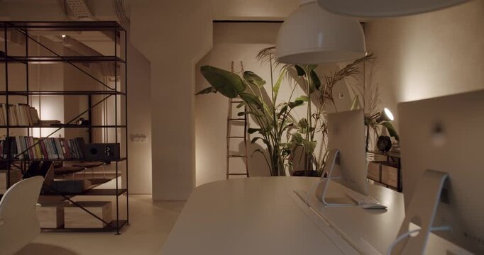 Empty coworking space with white furniture and computers in a modern office with no people. Minimalist open space interior with plants and warm lighting, creating a comfortable and calm atmosphere