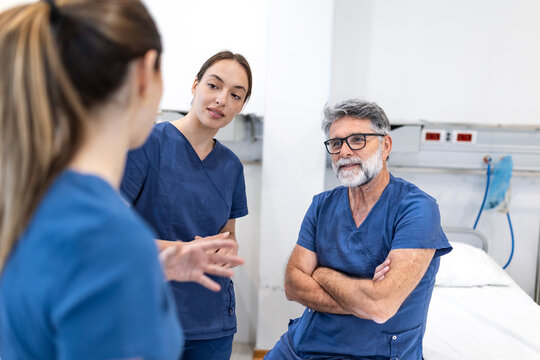 Senior male surgeon with glasses and grey hair sitting and talking with two young female medical professionals in a bright hospital room, reviewing patient information in a collaborative work setting.