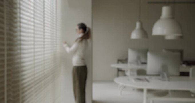 Slow motion footage of a female employee in a modern minimalist office walking to the window and peering outside through the venetian blinds, expressing curiosity and a need for a break
