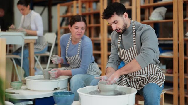 Man mold products on potters wheel and talk to ceramist girl. Relaxed working atmosphere, hobbies, master classes for adults and young people. 