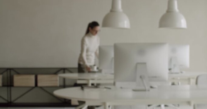 Determined businesswoman in a minimalist coworking space walking towards her desk with a coffee cup in hand, preparing to start her workday at the computer in a bright, modern interior