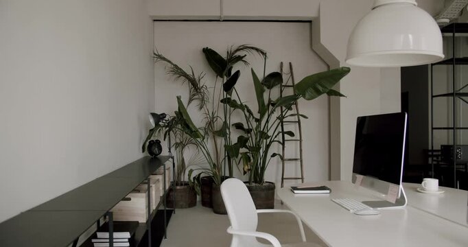 Modern and stylish office interior featuring a clean white desk, a computer, comfortable chair, and large green plants creating a biophilic design concept for a productive workspace