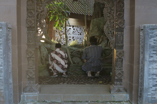 Two Indonesian men praying inside a traditional carved wooden gate with sacred banyan tree roots.