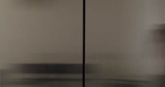 Mysterious silhouette of a person walking behind a textured and translucent fluted glass door, creating an abstract and distorted visual effect of movement and shadows in a modern interior