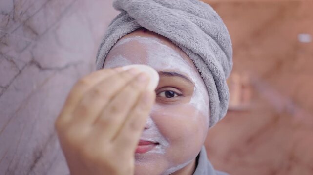 Young lady cleaning off face pack using a cotton pad in the bathroom, self care and skincare concept.