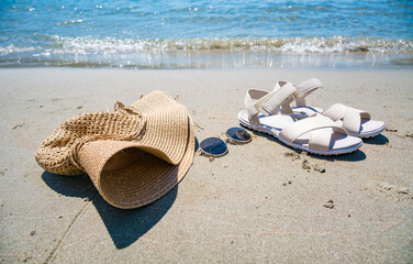A straw hat, sunglasses, and white sandals are placed on sandy beach near the water. The scene captures a relaxed summer vibe. © Mila