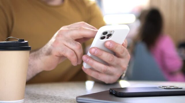 Person holds phone, taps screen while sipping coffee. Nearby laptop open on table. Background shows cafe with other patrons. Relaxed, focused mood during casual digital work or browsing