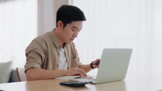 Happy young Asian man working on laptop in living room. Freelance worker male sitting at desk writing notes while watching webinar, studying online