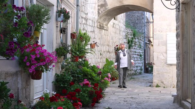 Mature Woman Walking in Flowered Old Town Street. Mature female tourist walking through a narrow stone street with flowers and old architecture. Peaceful sightseeing scene in a Mediterranean town.